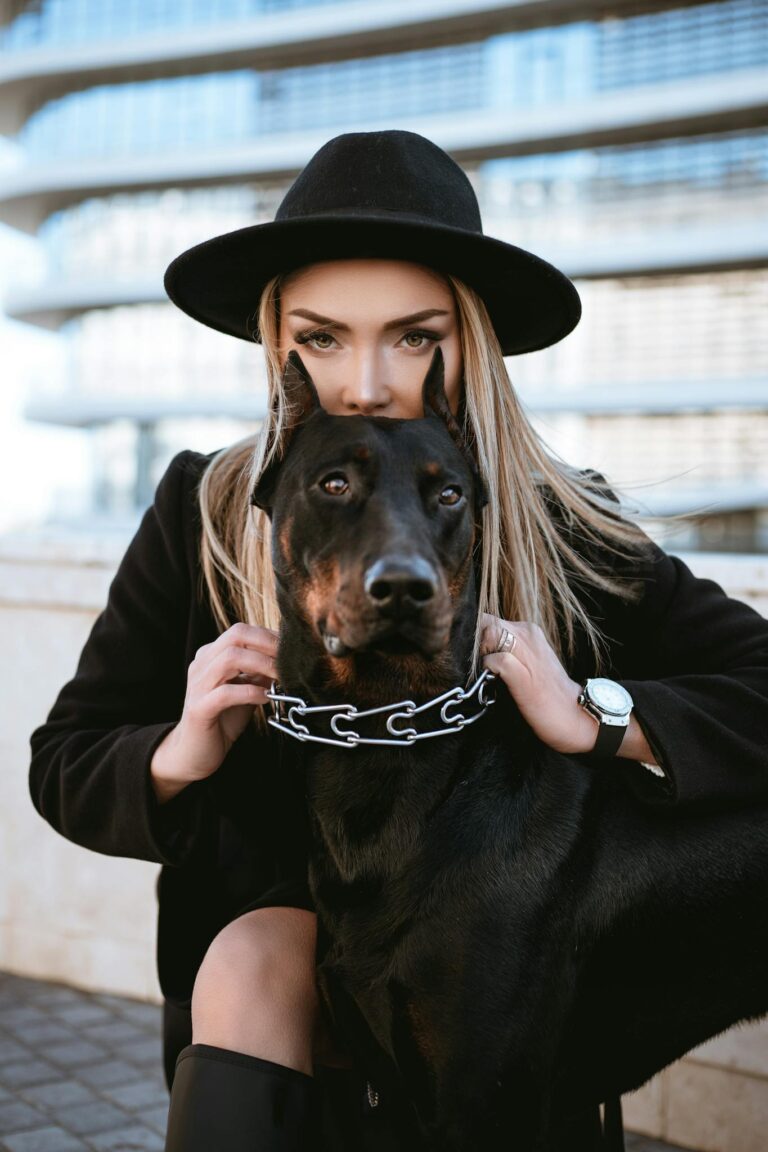 A fashionable woman poses with her Doberman outdoors against a modern urban backdrop.