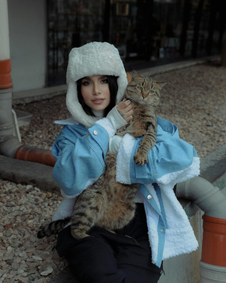 Stylish woman in a blue jacket and hat holding a tabby cat outdoors.